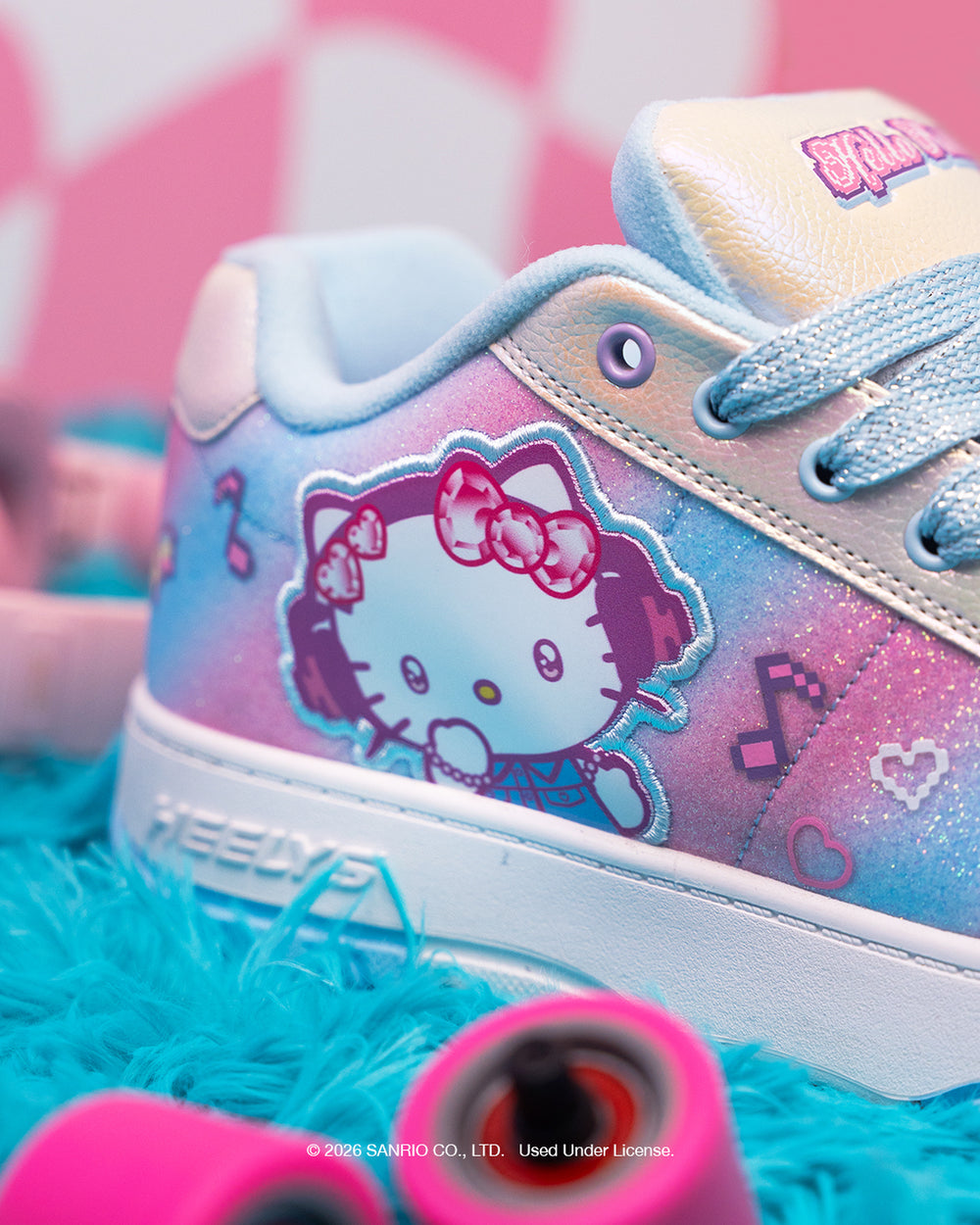 A close-up of a pastel Hello Kitty sneaker with pink and blue glittery panels, a large Hello Kitty graphic on the side, and light blue laces, styled on a turquoise fuzzy surface with pink skateboard wheels in the foreground.