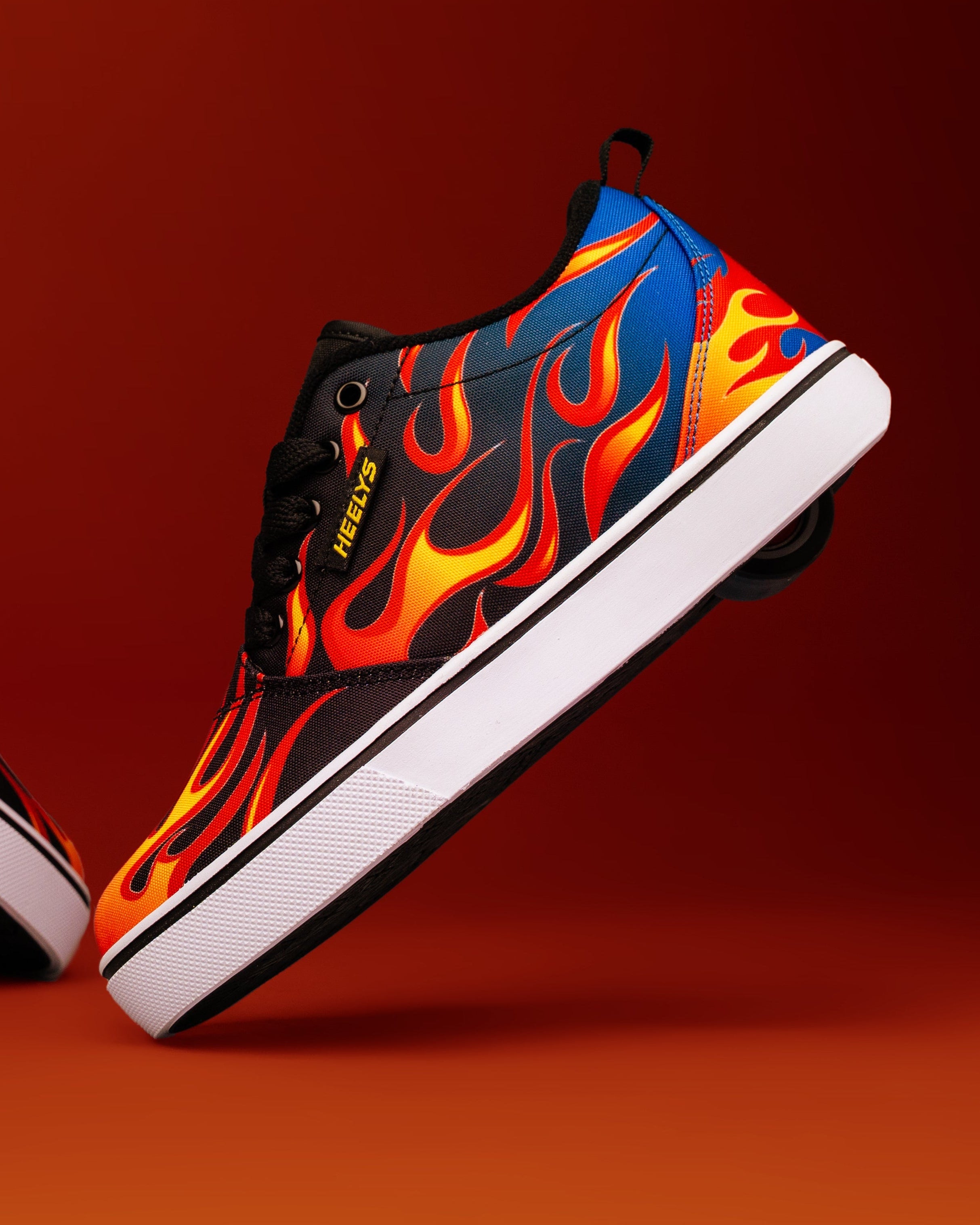 A pair of low-top sneakers with a black base and vibrant flame patterns in blue, orange, and yellow, set against a warm orange-brown gradient background.