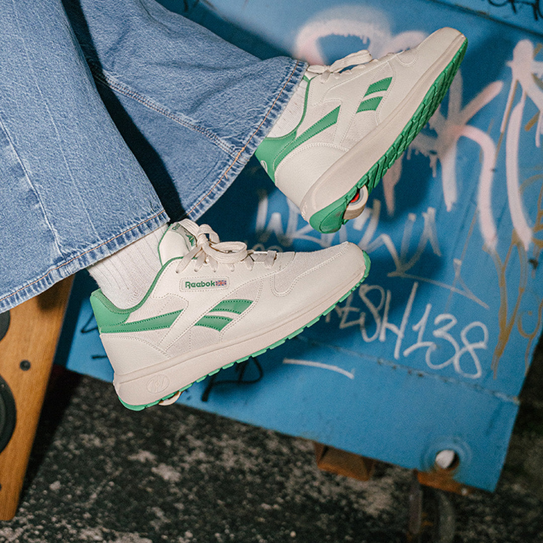 White and green sneakers with Reebok logo worn by a person sitting on a blue graffiti-covered surface.