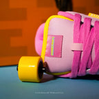 A close-up of a pink skate shoe with bright pink laces, a yellow wheel, and a pink heel counter, resting on a blue surface with an orange background.