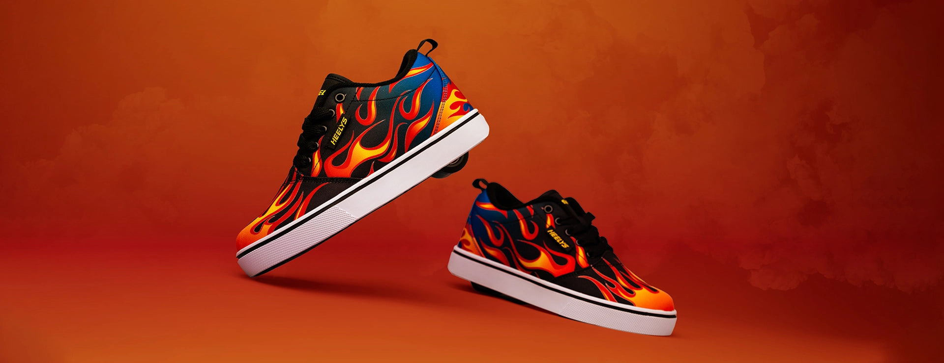 A pair of low-top sneakers with a vibrant orange, yellow, and blue flame pattern on a black base, featuring white midsoles, set against a warm orange gradient background.