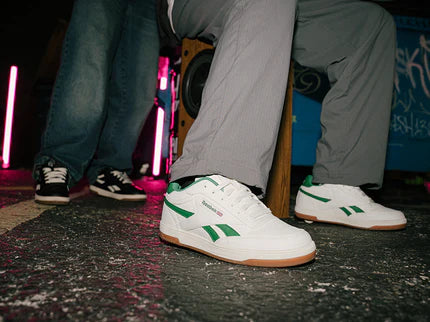 White sneakers with green accents worn by a person sitting on a stool.