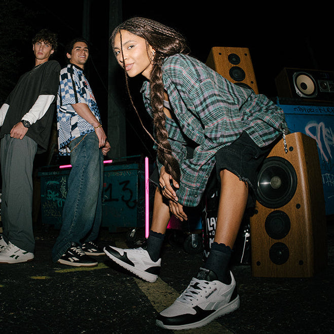 Person with braided hair posing in front of graffiti-covered walls and speakers.