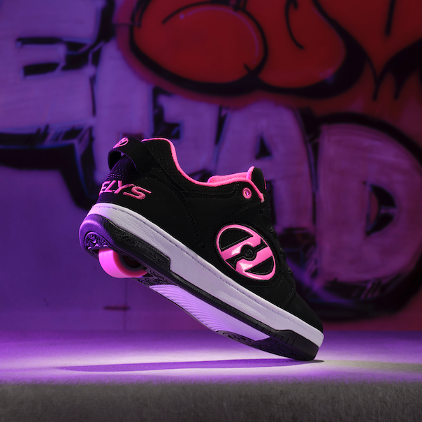 Black sneaker with pink Heelys logo accents on a dark surface with graffiti in the background
